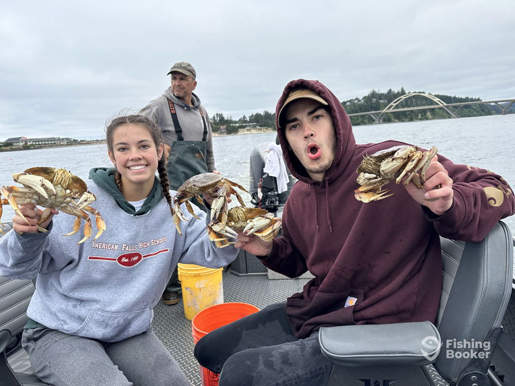 A young man and woman smile and pull faces while holding two large Crabs each near a bridge in Newport, Oregon with a fishing guide steering their boat in the background