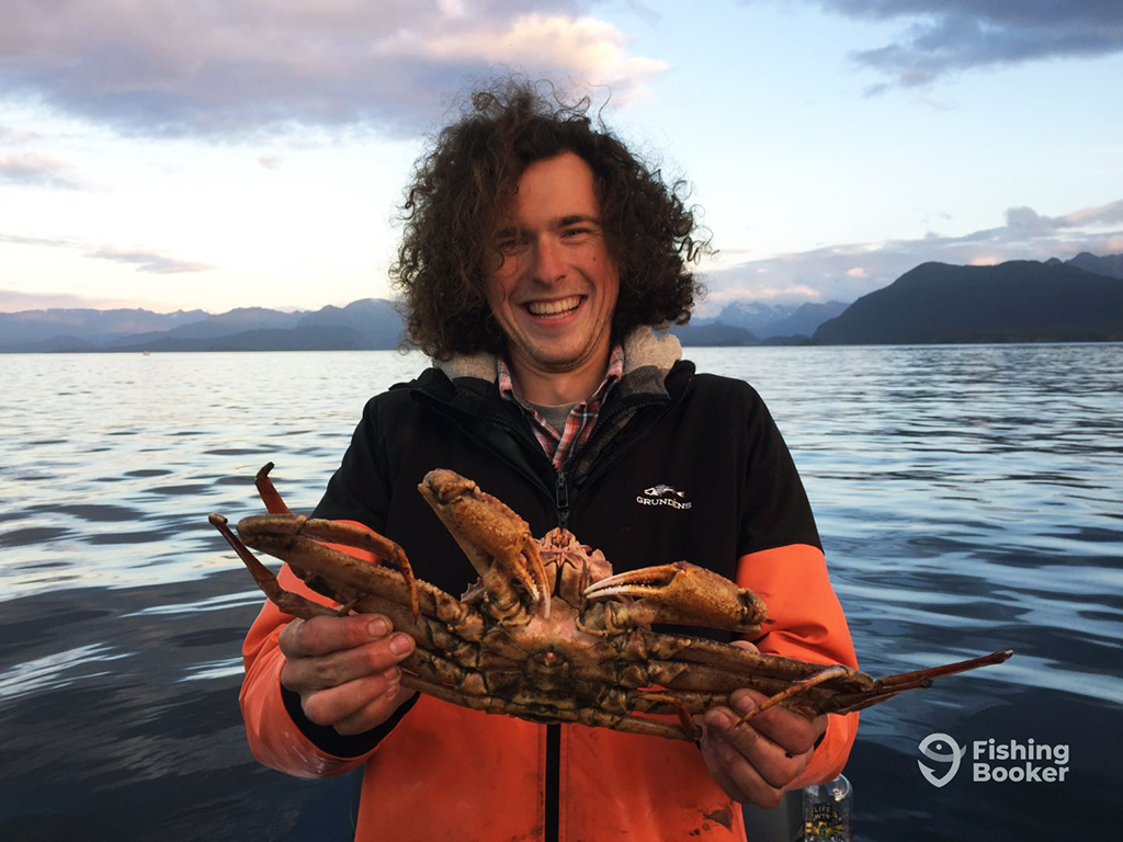 A man in a rain jacket holds a large Crab in both hands on a boat on a calm sea as the sun is setting