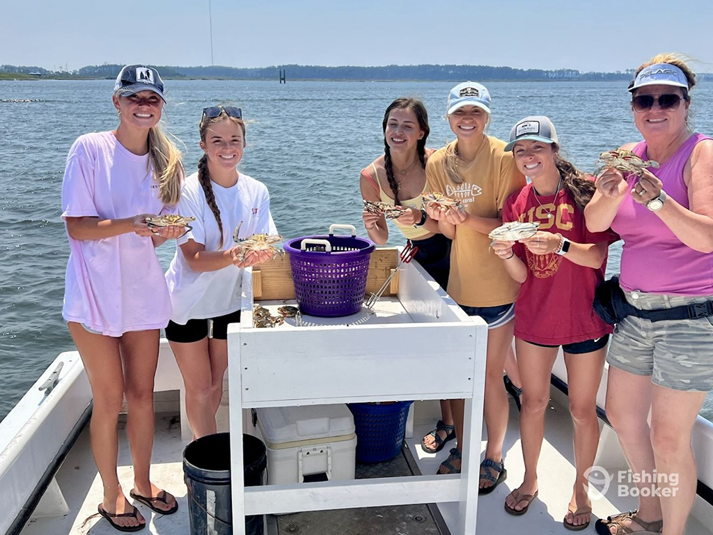 A group of young women hold a Crab each next to a purple Crabbing bucket aboard a large boat on a sunny day