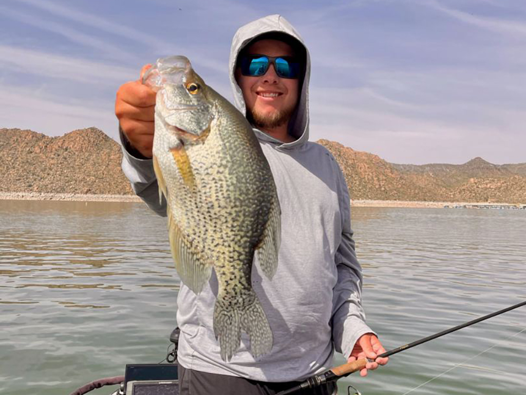 A happy angler holds a large Black Crappie towards the camera in one hand and a fishing rod in the other hand on a boat on a lake in Arizona
