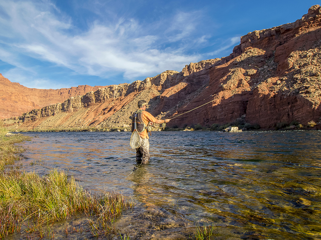 A lone fly fisher casts into shallow water in a river among rocky, dry mountains in Arizona on a clear day