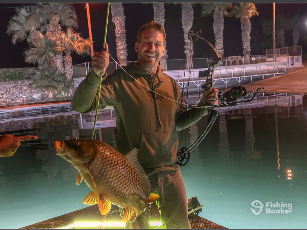 An angler holds a bow and a bowfishing arrow that's speared into a Carp on a boat close to shore at night, with palm trees visible on the shore behind him