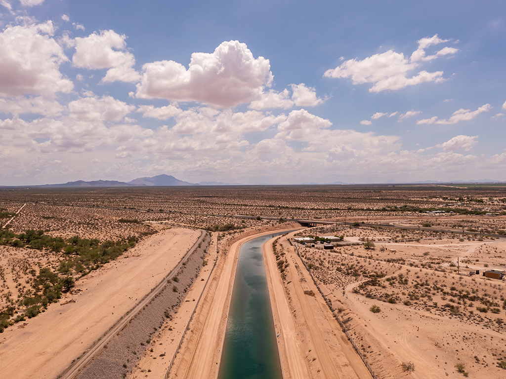 An aerial view of an irrigation canal running through the middle of a sandy desert plain in Arizona with mountains in the background