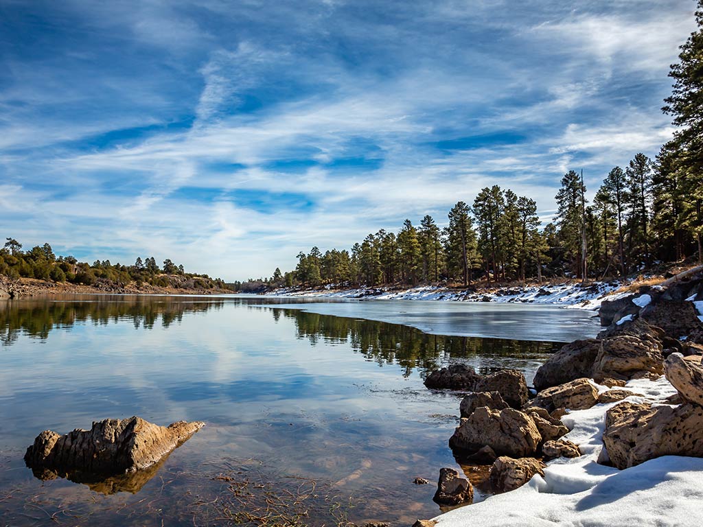 A view across the water from a snow-covered shoreline of a lake in the White Mountains in Arizona on a winter's day with sunny intervals