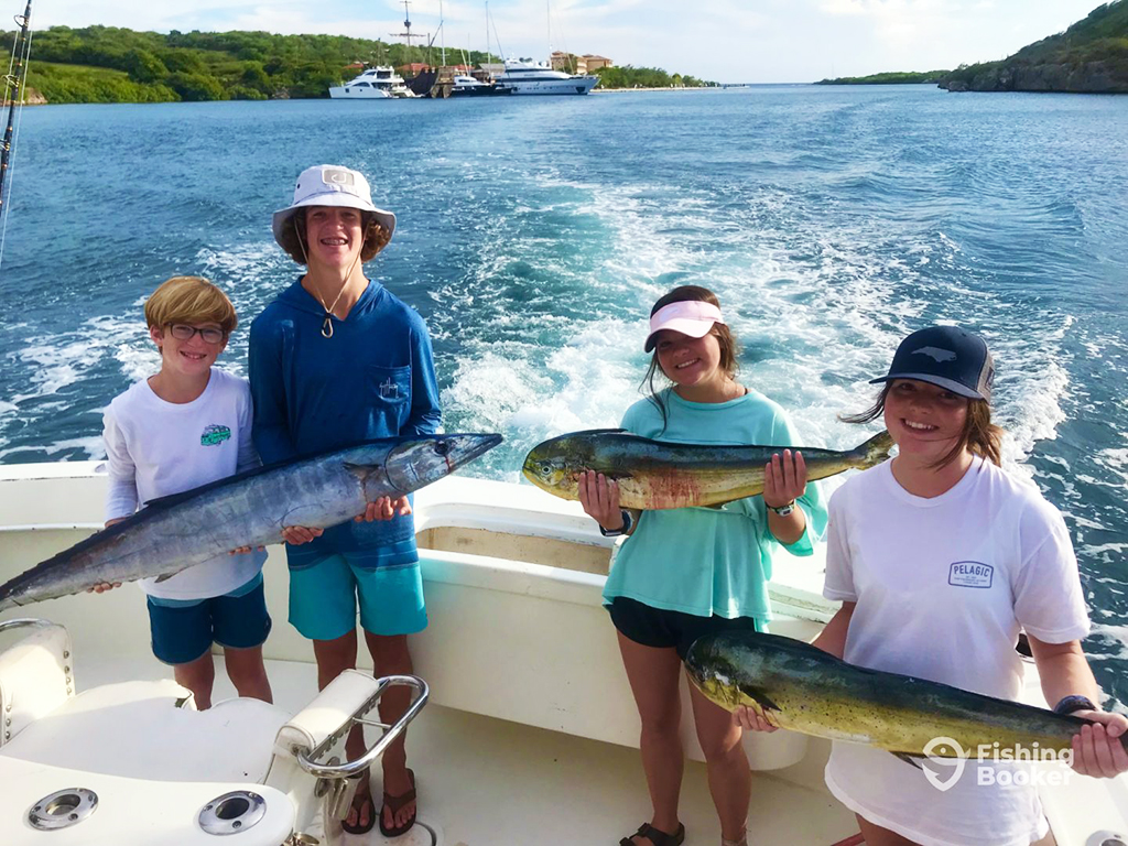 Four children stand aboard a boat holding two Mahi Mahi and one Wahoo near forested shorelines with a small group of yachts in the background