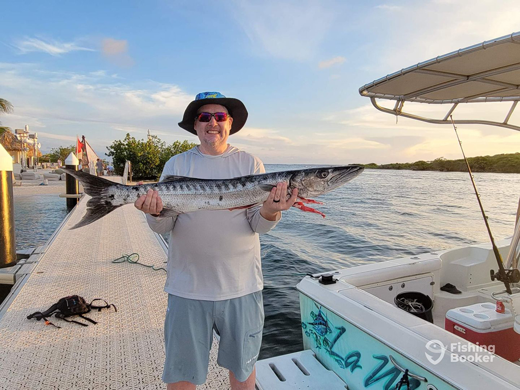 A pleased man holds a Barracuda while standing on a Curaçao dock next to a boat on a sunny day