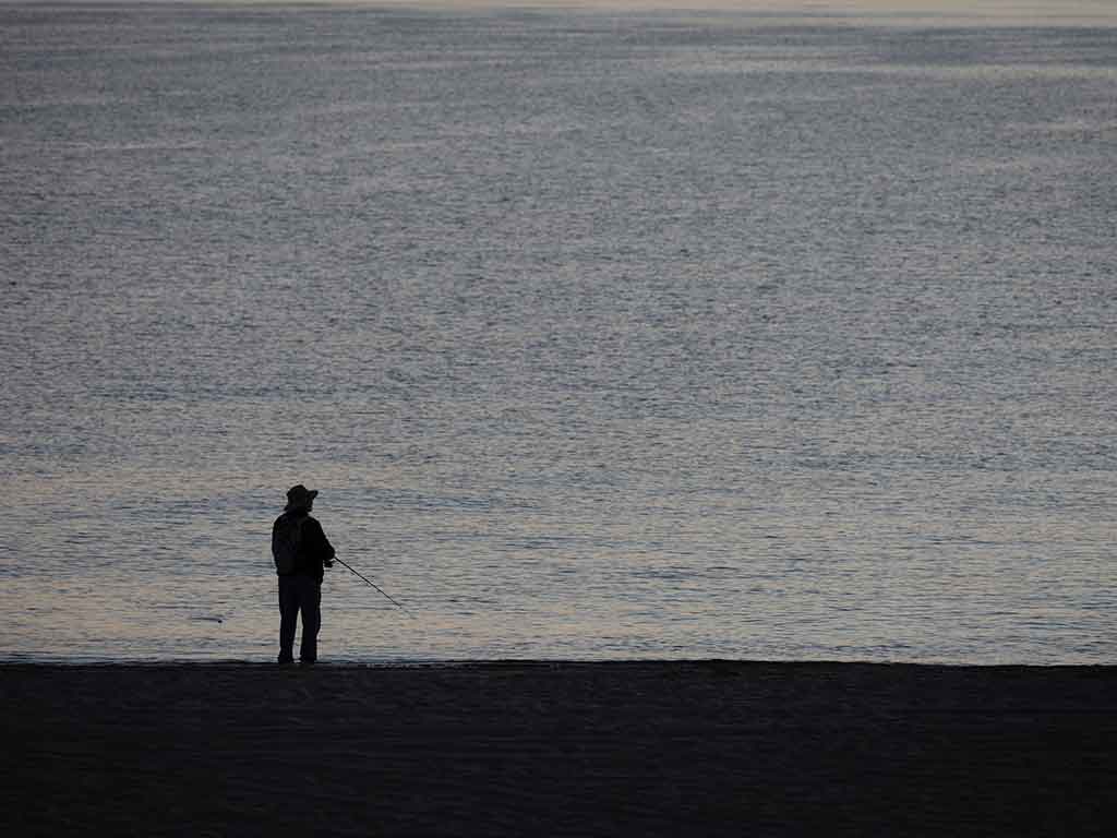 A view from behind of a lone angler fishing from shore as it's getting dark, effectively making the angler a silhouette against a background of the water