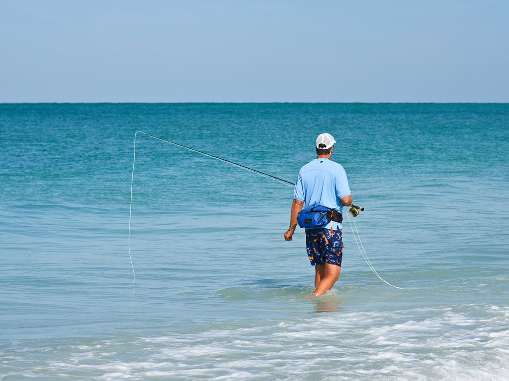 An angler wades through shallow water close to shore holding a fly rod ready to go fly fishing