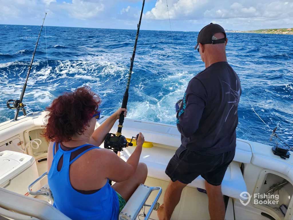 A woman sits in a fighting chair while deep sea fishing with heavy tackle next to a charter boat captain on a boat in Curaçao on a clear day 