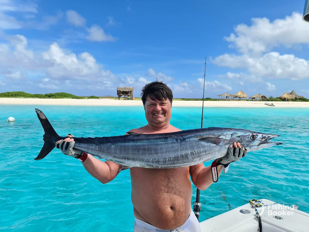 A happy angler holds a large Wahoo on a boat by clear blue seas and a white sandy shoreline on a sunny summer day