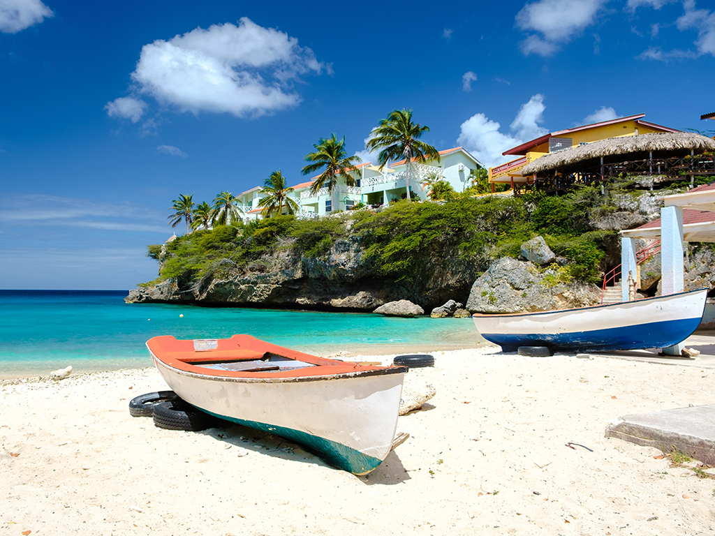 Two small wooden fishing boats sit on a sandy beach in Curacao next to the calm green-blue waters of a bay and in the shadow of a small palm tree-lined cliff with houses on it