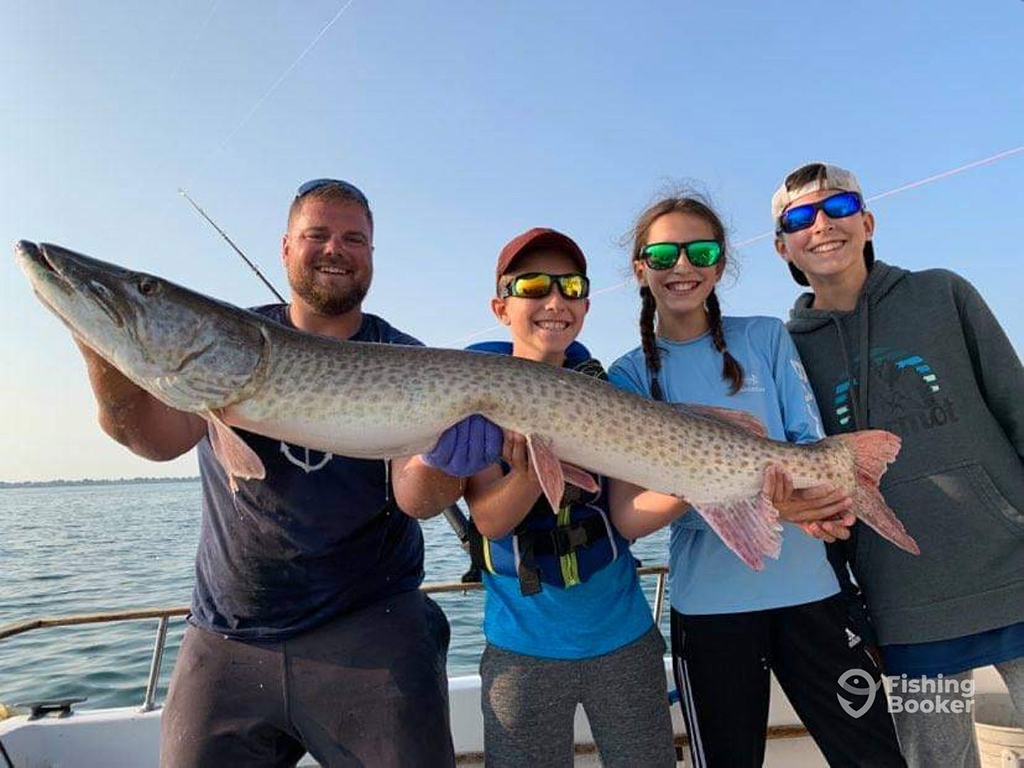 A family consisting of a man and three children holding a large Musky between on a boat on a sunny day