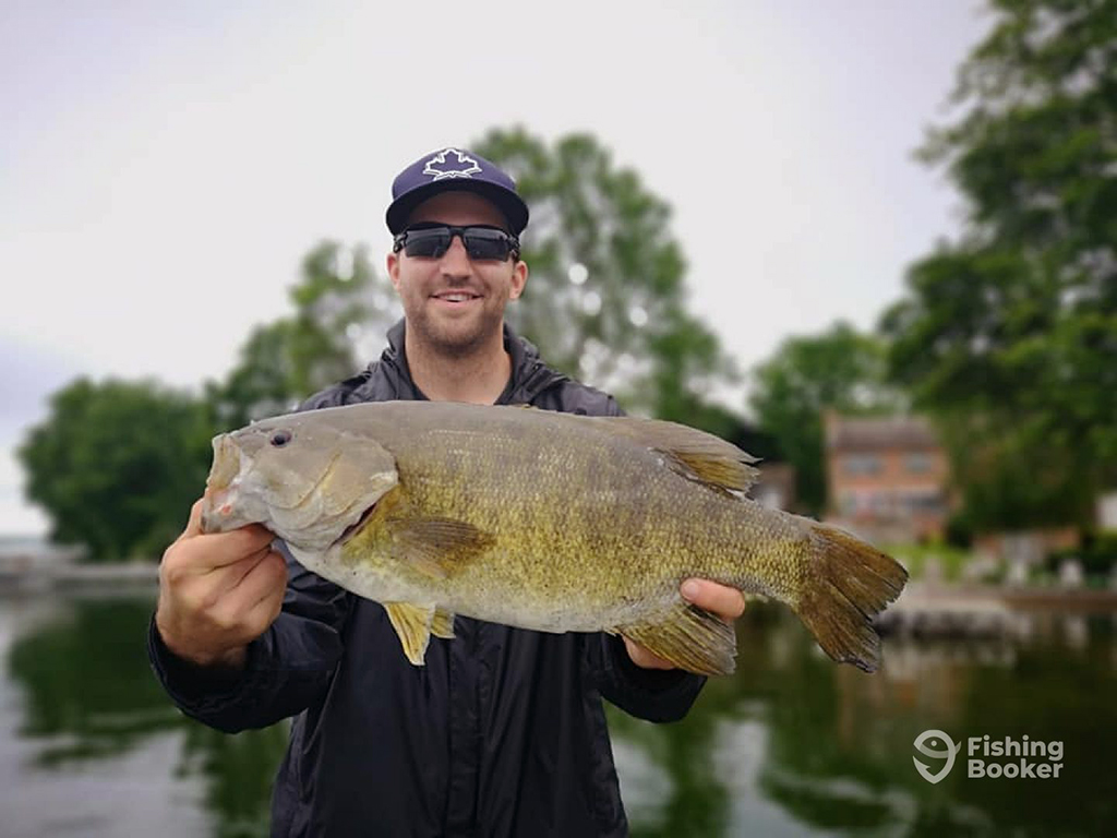 A man in a baseball cap and sunglasses holds a large Smallmouth Bass on a river on a cloudy day