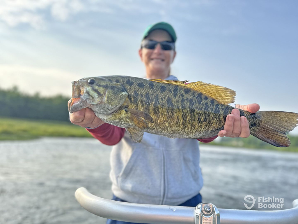 A woman holds a Smallmouth Bass towards the camera while standing aboard a boat on a lake on a sunny day