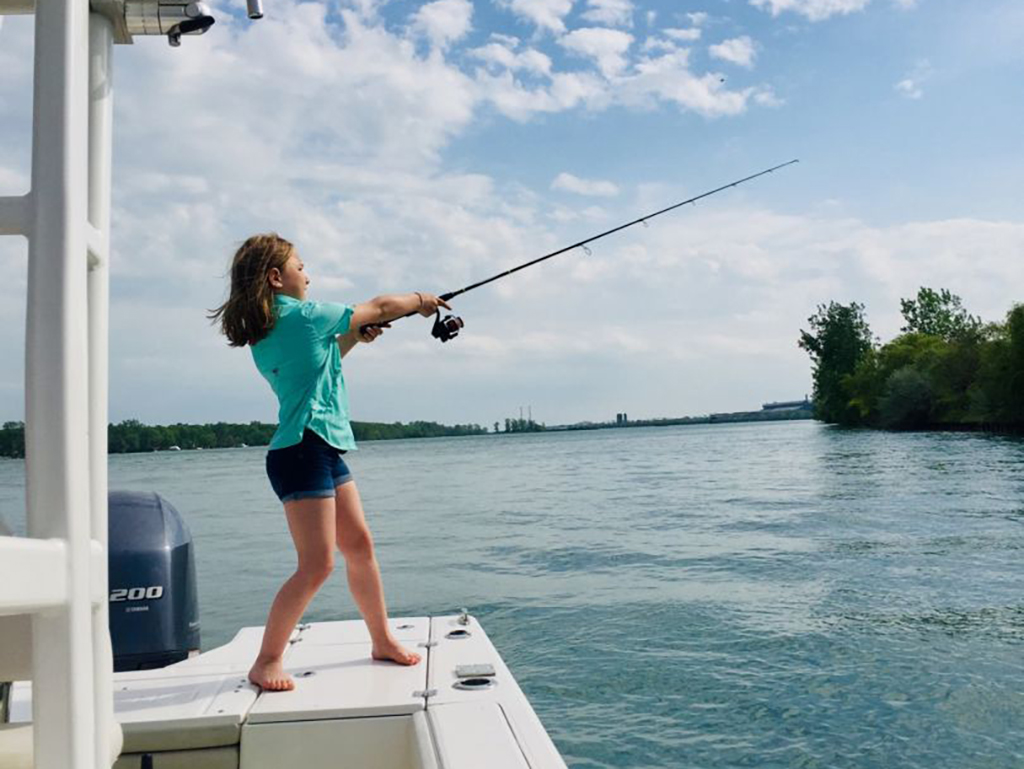 A view along the side of a boat of a young girl casting for a Smallmouth Bass from a boat on a lake on a sunny day