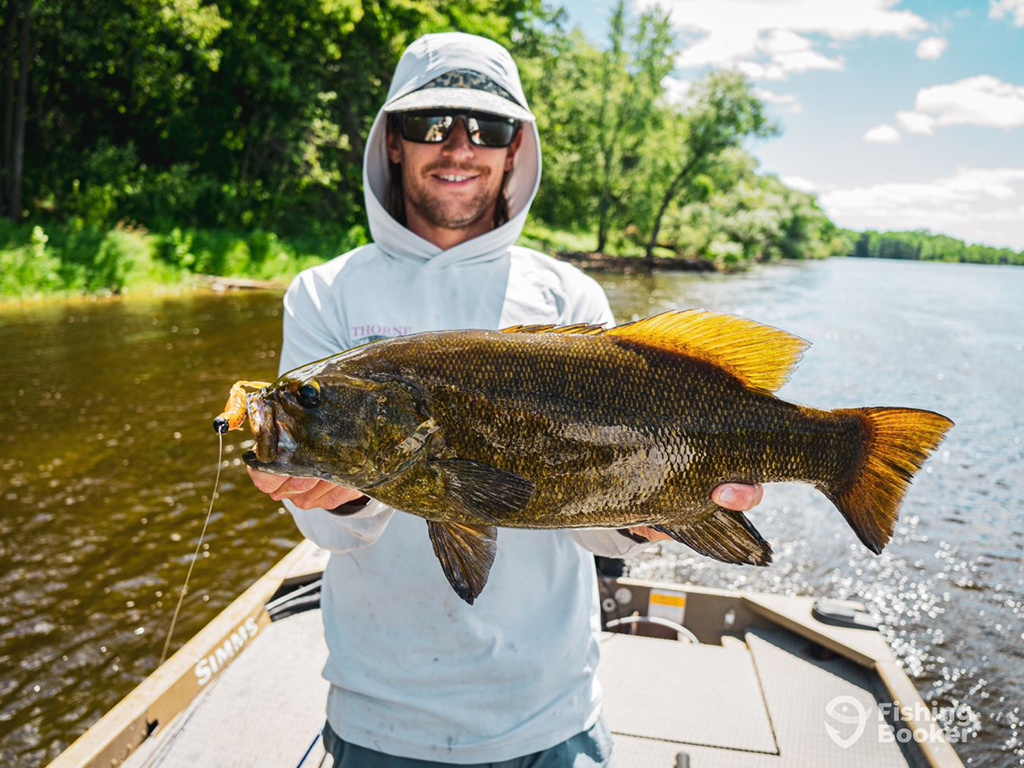 A happy angler on a boat on a shallow river holds a large Smallmouth Bass that still has the artificial lure in its mouth that was used to catch it