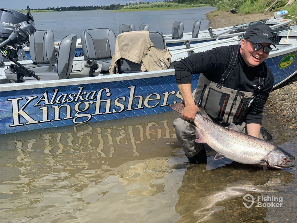 A smiling angler wearing waders crouches in shallow river water while releasing a Chinook Salmon near a river bank next to an Oregon fishing guide's boat