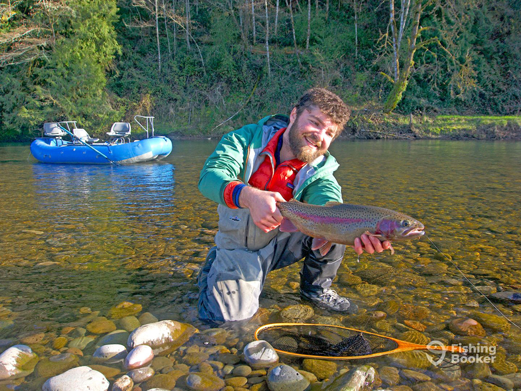 An angler kneels in a shallow river as he holds a Cutthroat Trout prior to releasing in with his inflatable drift boat in the background