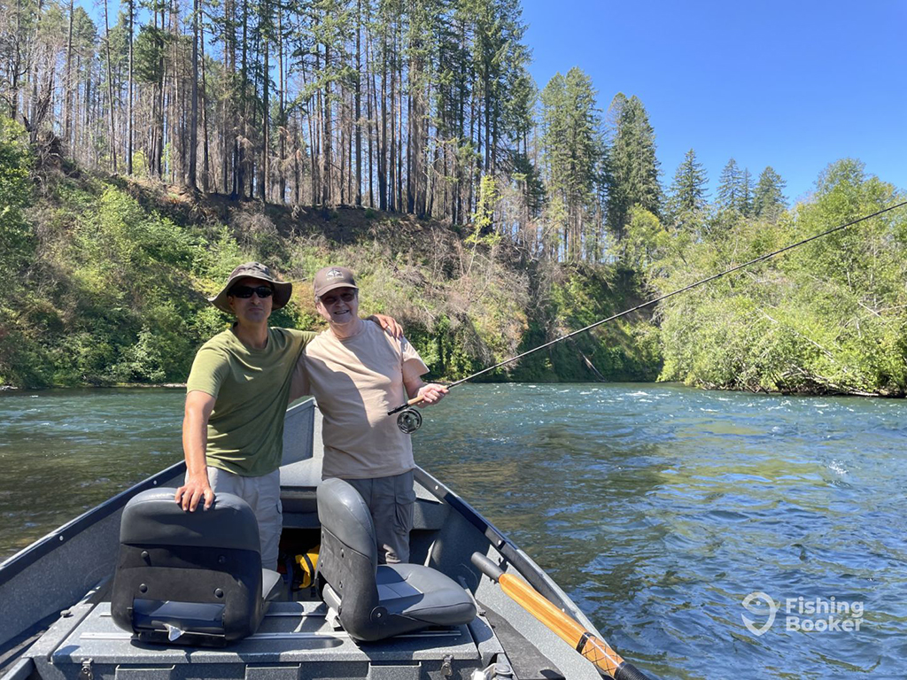 Two anglers stand aboard a row boat holding a fly fishing rod on a river near Eugene, Oregon against a backdrop of early spring foliage 