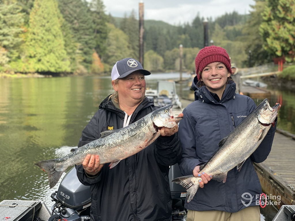 Two smiling female anglers in hats and rain jackets smile as they hold a Salmon each on a river near a dock in the early fall