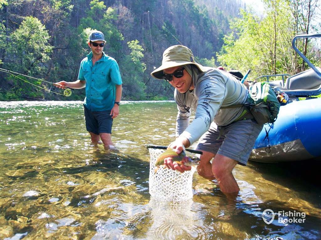 A man stands in a shallow river holding a fly fishing rod while a fly fishing guide holds a Trout towards the camera in one hand and a release net in the other. Their drift boat is visible to the right of the photo