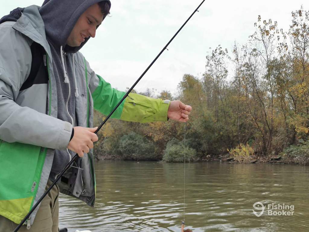 An angler in a hooded sweatshirt holds a light fishing rod in one hand and a fishing line in the other against a wintery river backdrop