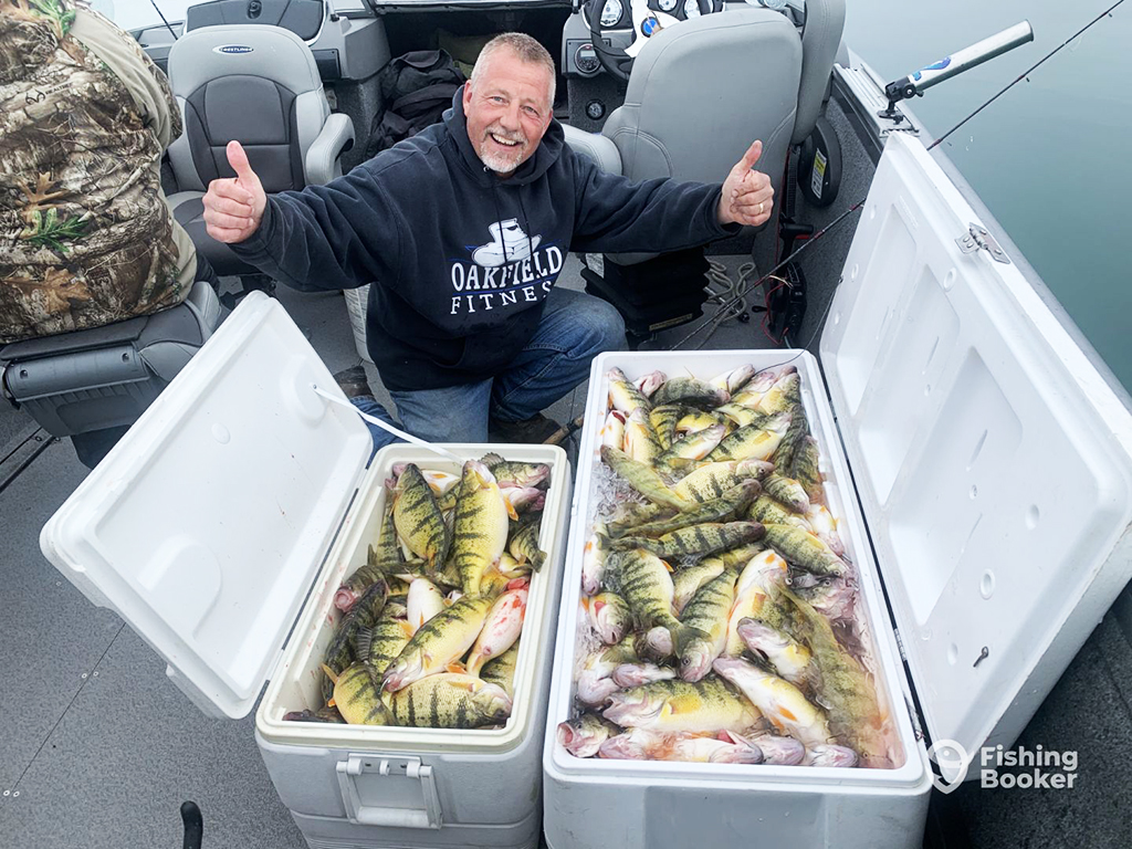 A happy angler kneels on a boat showing two thumbs up next to two large ice chests full to the brim of Yellow Perch 