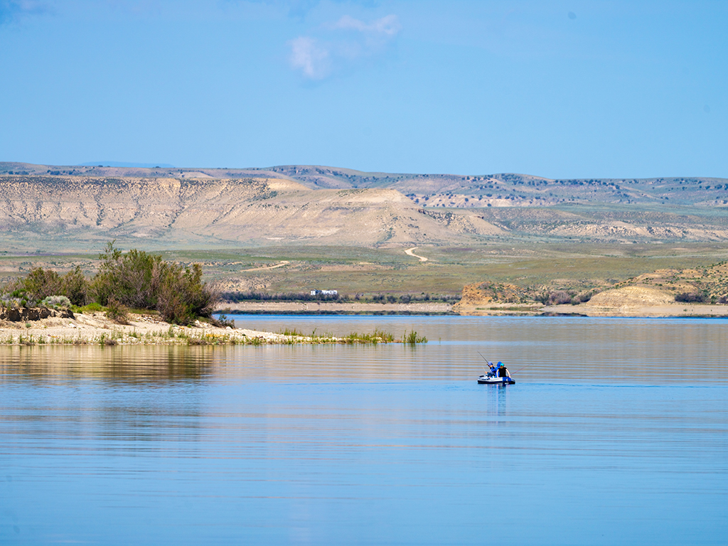 Two anglers fish from a small boat in the distance on a large reservoir in Utah on a sunny day, with yellow, grass-covered hills in the background