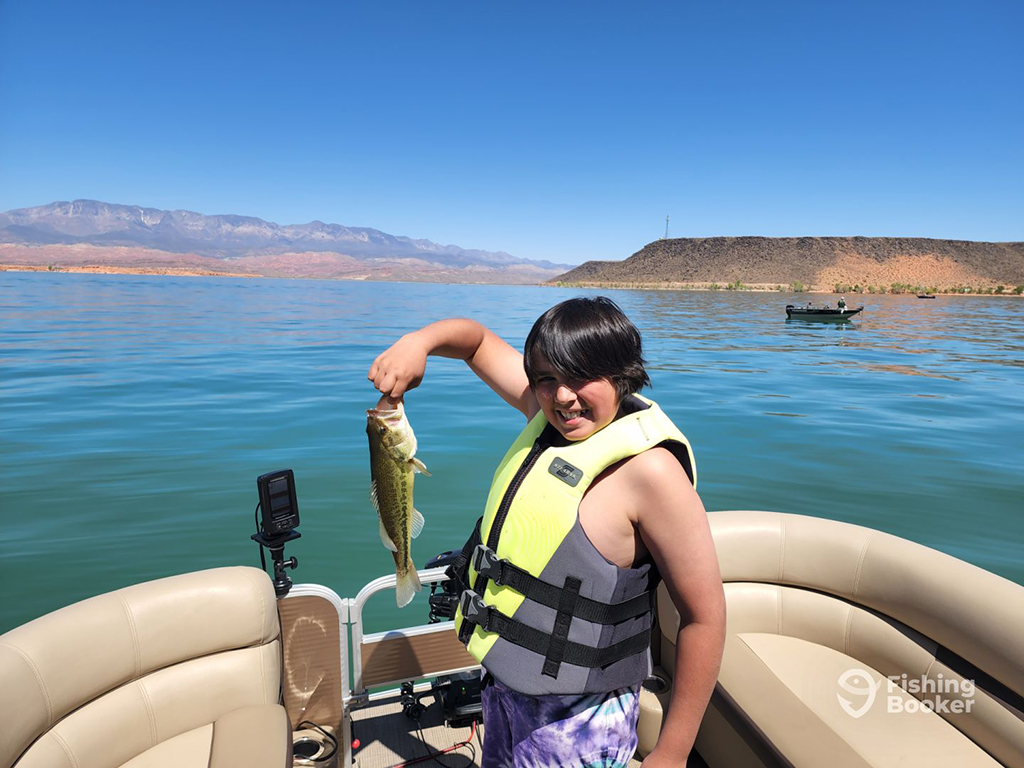A young angler in a personal floatation device holds a small Largemouth Bass aboard a pontoon boat on a lake in Utah on a sunny day