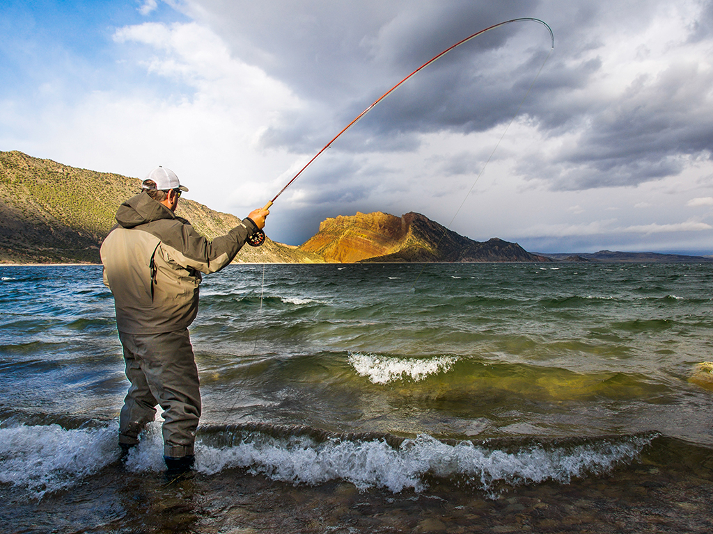 An angler stands in shallow water with his back towards the camera as he fights a fish with fly fishing equipment against a background of dramatic yellow rocks and stormy skies in the Flaming Gorge Nation Recreation Area, Utah