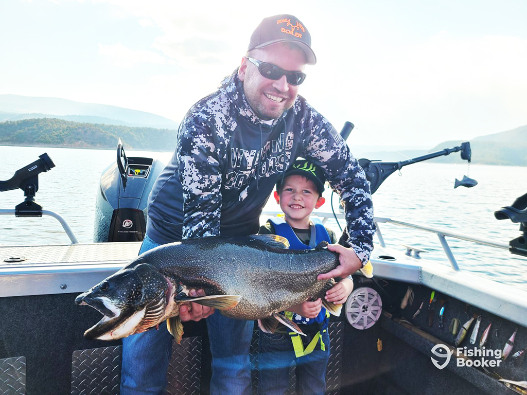 A charter captain helps a young boy hold a large Lake Trout on a boat on a lake in Utah. Downriggers and other fishing equipment are visible in the background
