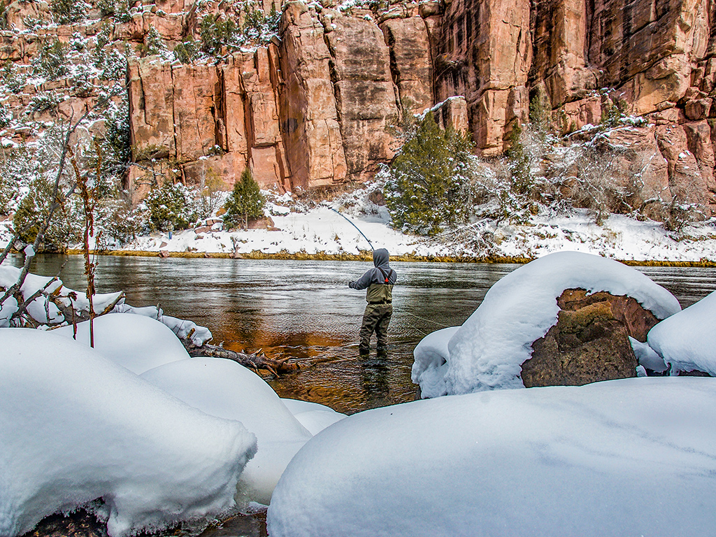 An angler wearing waders and a hooded sweatshirt fly fishes in a canyon on the Green River in the Flaming Gorge National Recreation area, Utah, on a snowy, sunny winter's day