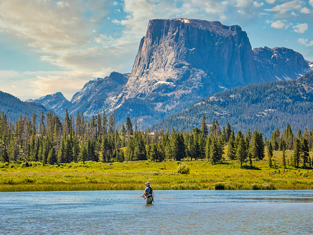 A solitary man stands in a shallow lake in Wyoming while using fly fishing equipment, with tall rocky outcrops and pine trees visible in the background