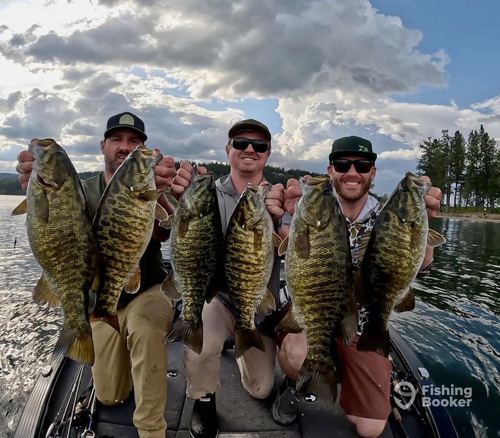 Three smiling male anglers each holding two large Smallmouth Bass towards the camera as they kneel aboard a small boat on a lake