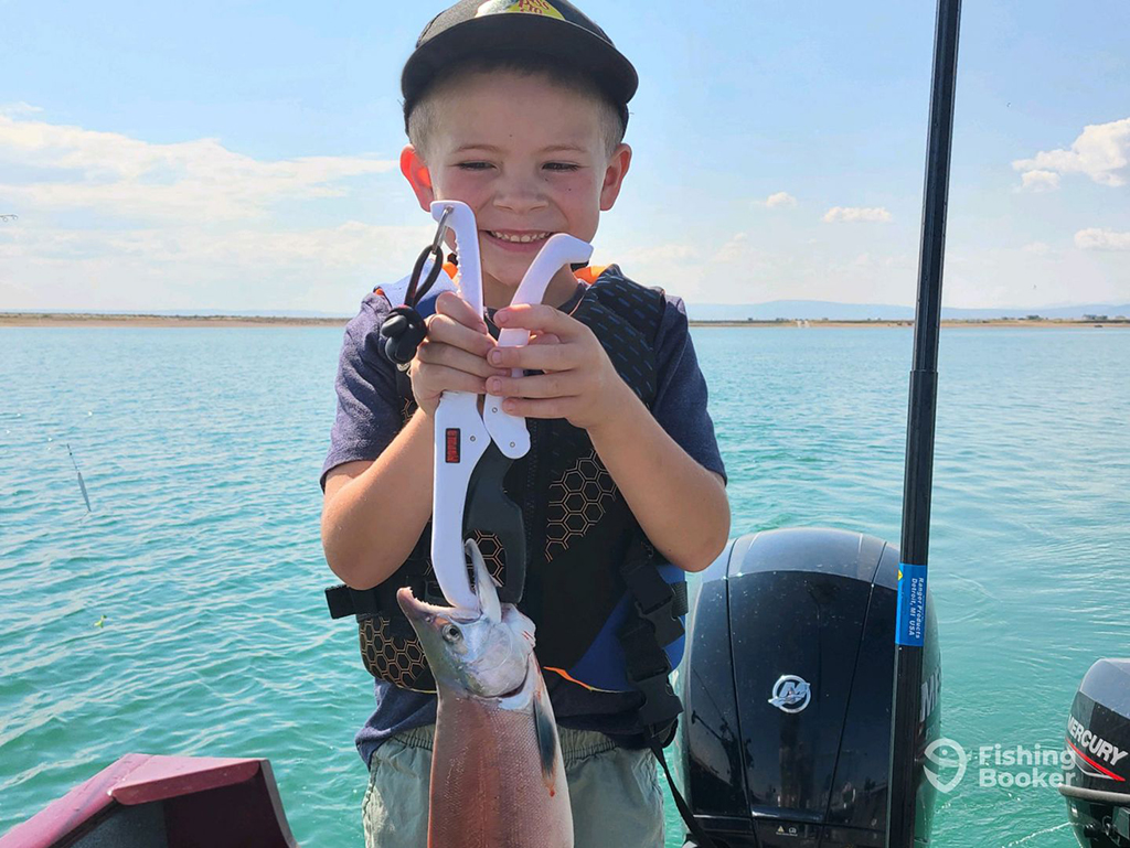 A young boy smiles as he holds a Kokanee Salmon on a lake in Wyoming on a clear blue sunny day