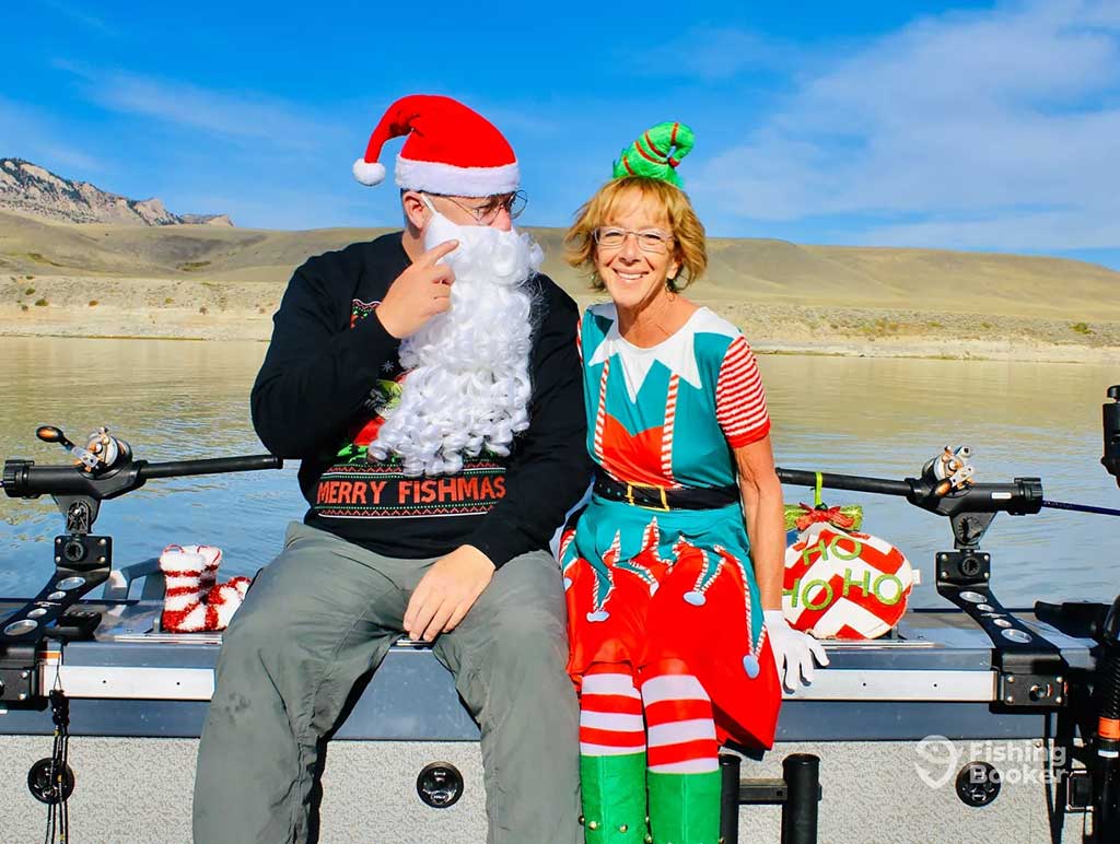 A man and a woman sit and laugh on a boat on a lake on a sunny day. They are wearing Christmas outfits including a sweater that reads "Merry Fishmas" with fishing reels and holiday-themed amenities visible around them