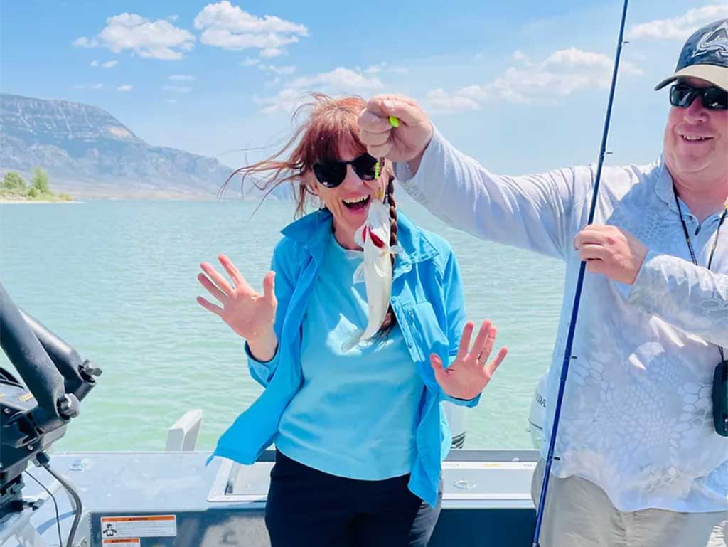 A man to the right of the photo holds a fishing rod in one hand and a small fish in the other, while a laughing woman in the center of the image holds her hands open, away from the fish that is being dangled in front of her. A beautiful mountain view and clear blue lake waters are visible behind the pair, who are standing aboard a boat.