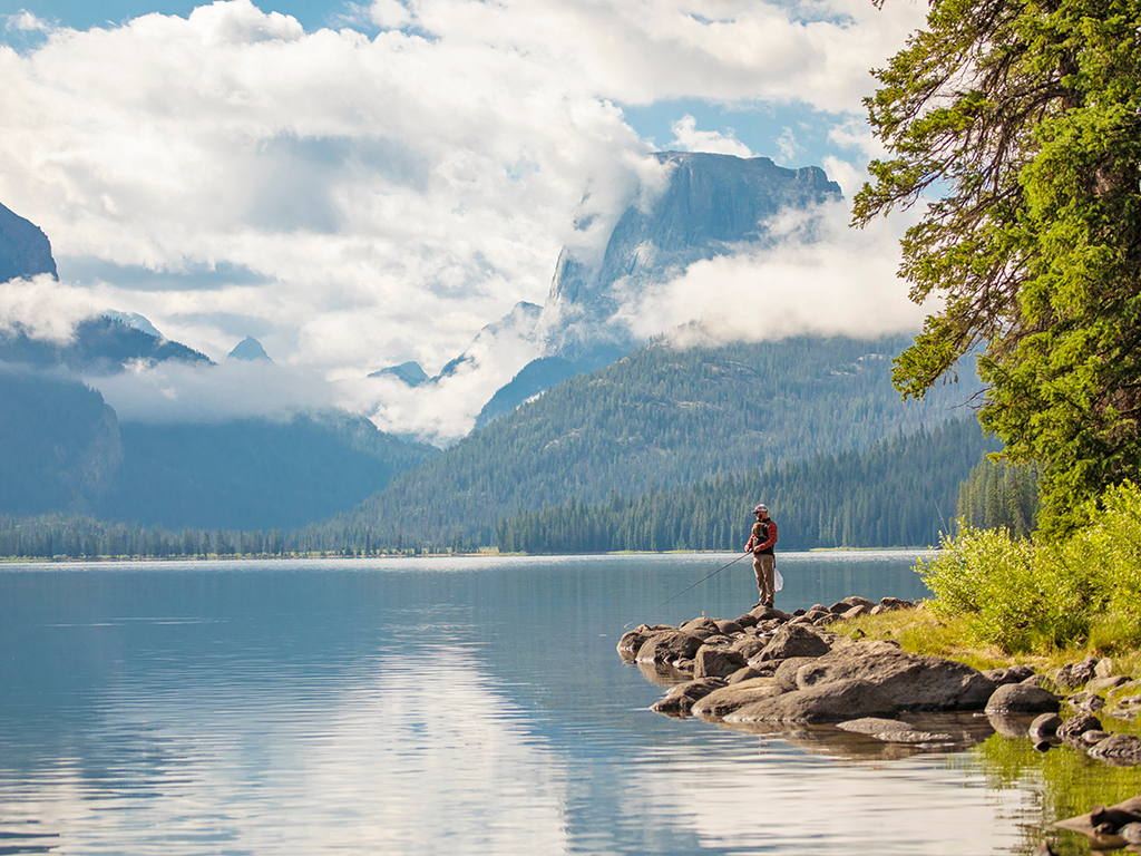 A man stands on a rocky shoreline while holding a fishing rod and fishing a calm Wyoming lake in sunshine and patchy, low clouds against a dramatic rocky backdrop