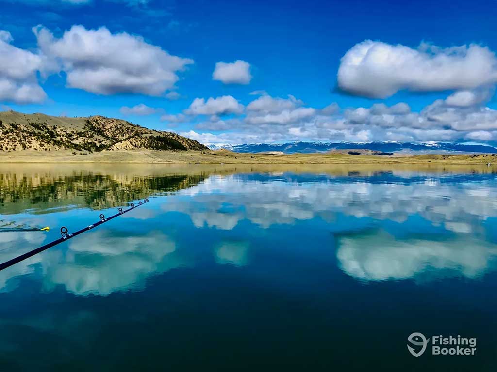 A fishing rod stretches out across a deep blue lake that's reflecting the blue, slightly cloudy sky and mountainous Wyoming shoreline