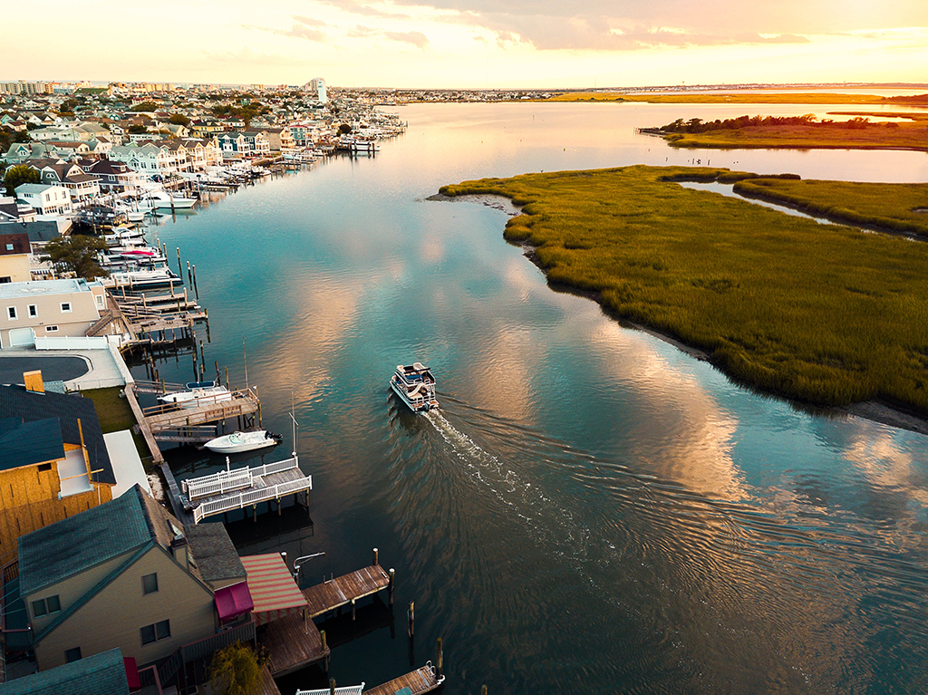 An aerial view of a boat traveling along a bay at dawn with the shoreline of Wildwood, NJ to the left with boat docks and houses, and grassy wetland broken up by rivers to the right