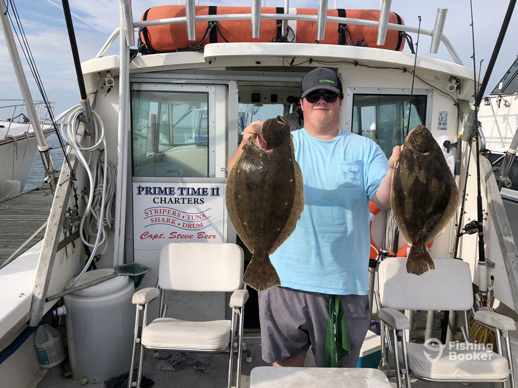 An angler holds up two large Flounder while standing aboard a large charter fishing boat in Wildwood, NJ on a day with sunny intervals