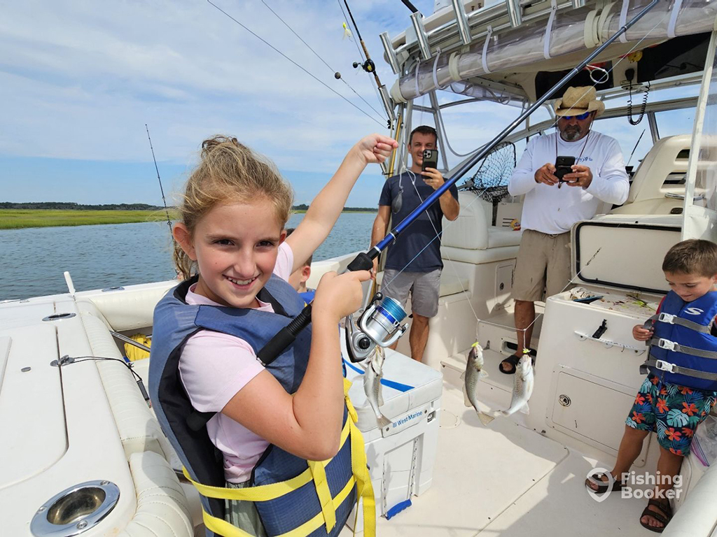 A child in a floatation device smiles as she holds a fishing rod and a line that has three small fish attached, whlie standing on the deck of a large charter boat in Wildwood, NJ, with a younger child and two adults behind her