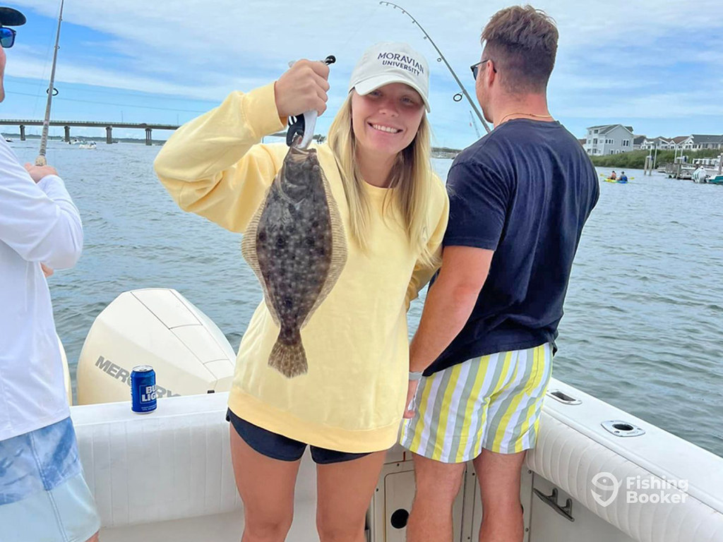 Two anglers stand aboard a boat near a bridge close to Wildwood, NJ. A woman smiles as she holds a Fluke while a man faces the water while holding a fishing rod