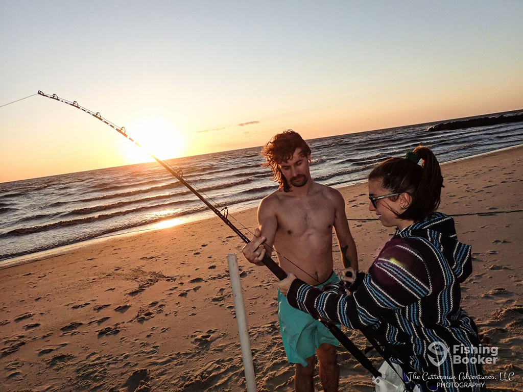 A fishing guide tutors a woman who is holding a large surf casting rod while standing on a beach in New Jersey as the sun is rising