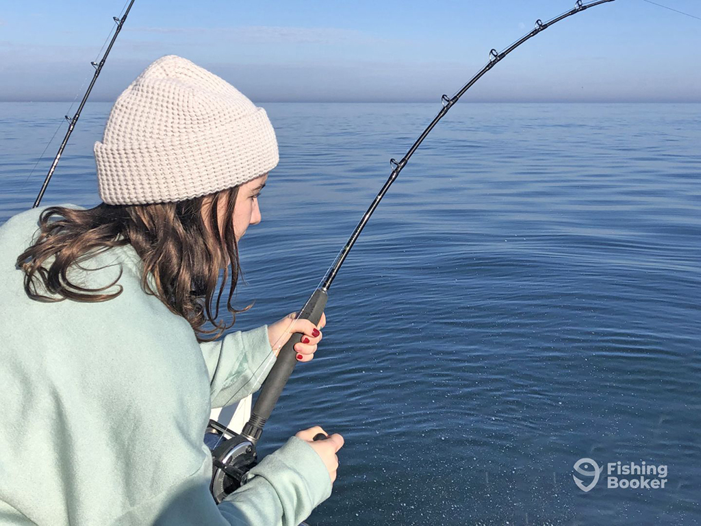 A girl in a hat leans over the side of a boat while reeling in a heavy fishing rod in a calm blue sea