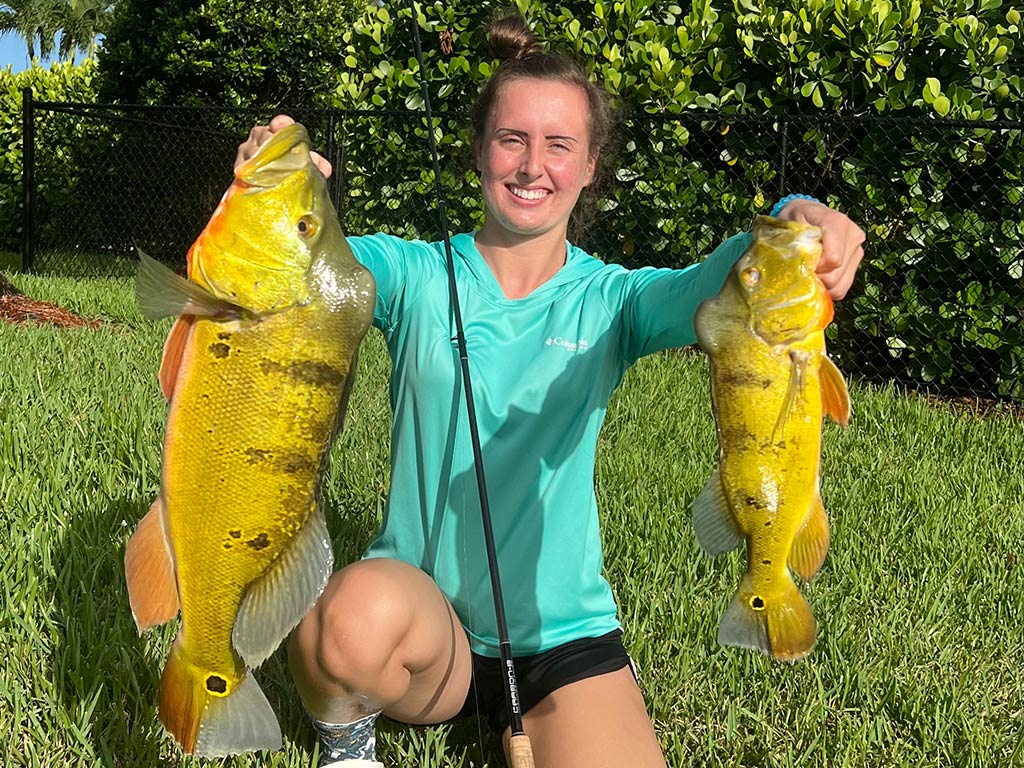 A woman kneeling on grass after a successful fishing trip in Miami, holding a Peacock Bass in each hand on a clear day