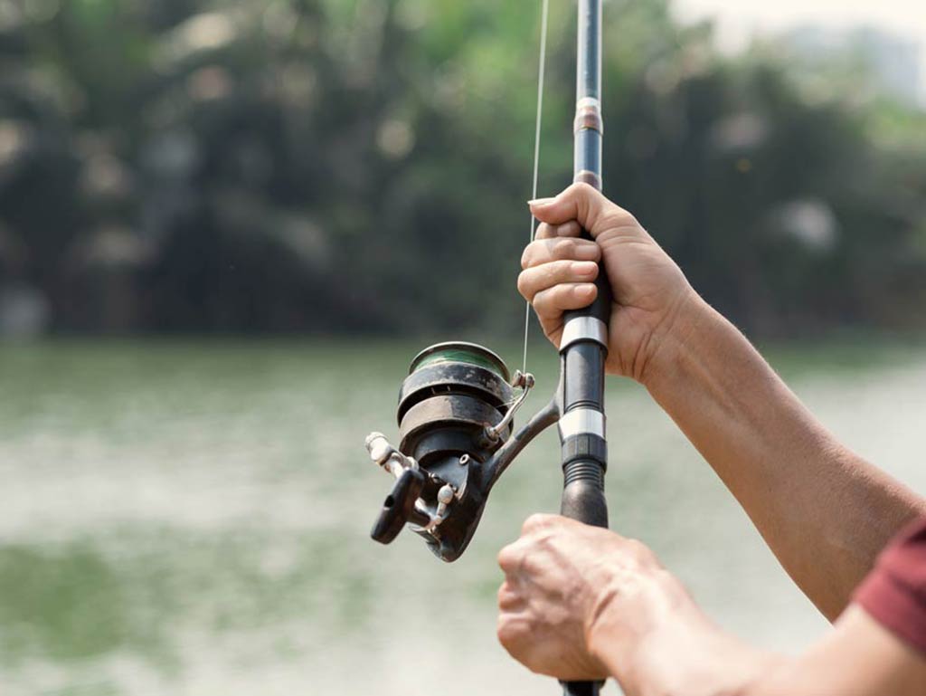 A close-up of a fishing rod and reel being held by an angler, with water and greenery blurred in the background