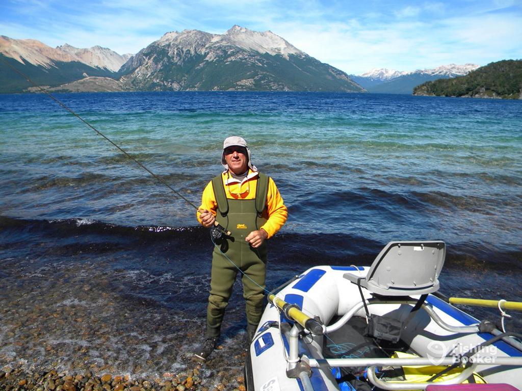 An angler wearing waders stands next to an inflatable boat and holds a fly fishing rod on the shore of a glacial lake with mountains in the background in Patagonia, Argentina