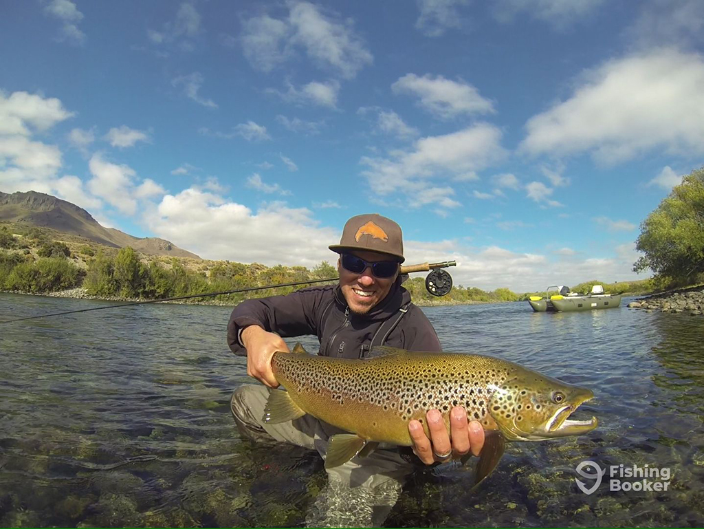 An angler kneels in shallow water near the shore of a lake while holding a large Brown Trout and a fly fishing rod. An inflatable boat is visible behind him, as well as mountainous scenery and green trees in the background