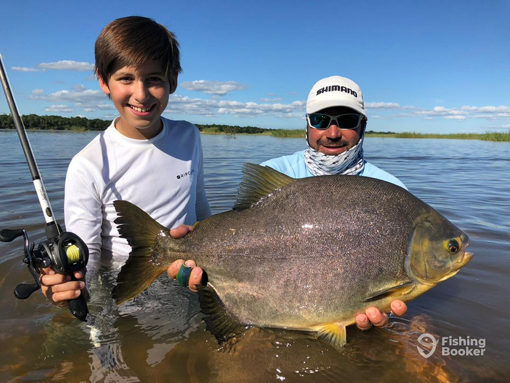 A teenage boy holding a rod with a baitcaster reel stands next to an Argentina fishing guide who's kneeling in a muddy river holding a large Pacu just above the surface